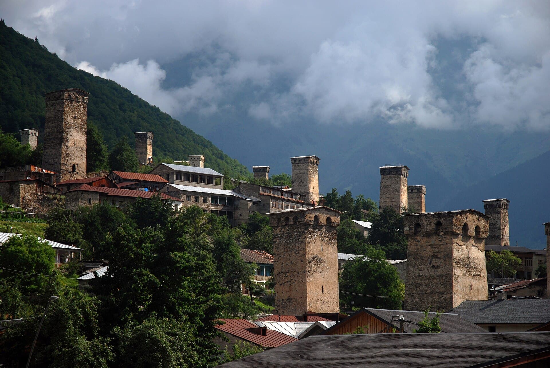 Die Steintürme von Mestia zwischen Häusern und Bäumen, Berge unter Wolken.