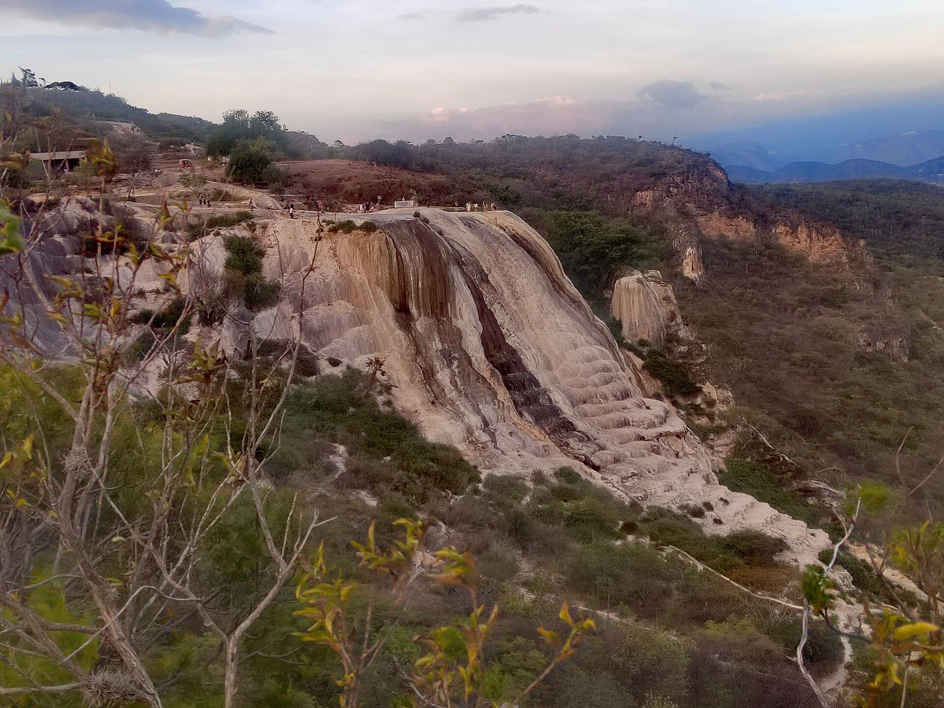 Weiße Gesteinsformationen fließen bei Hierve el Agua wie ein erstarrter Wasserfall eine Klippe hinab, umgeben von bewaldeten Bergen.
