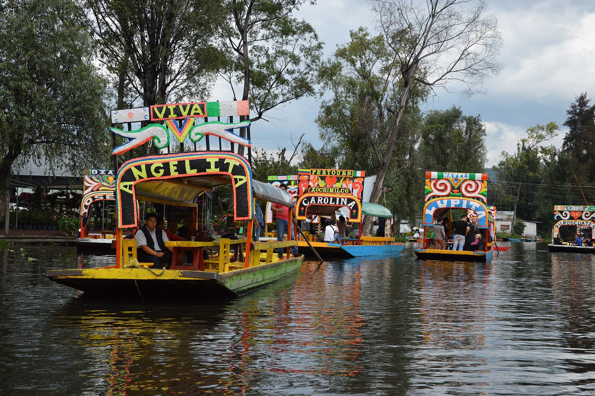 Brightly painted boats with names on their arches lined up on a tree-shaded canal in Xochimilco.