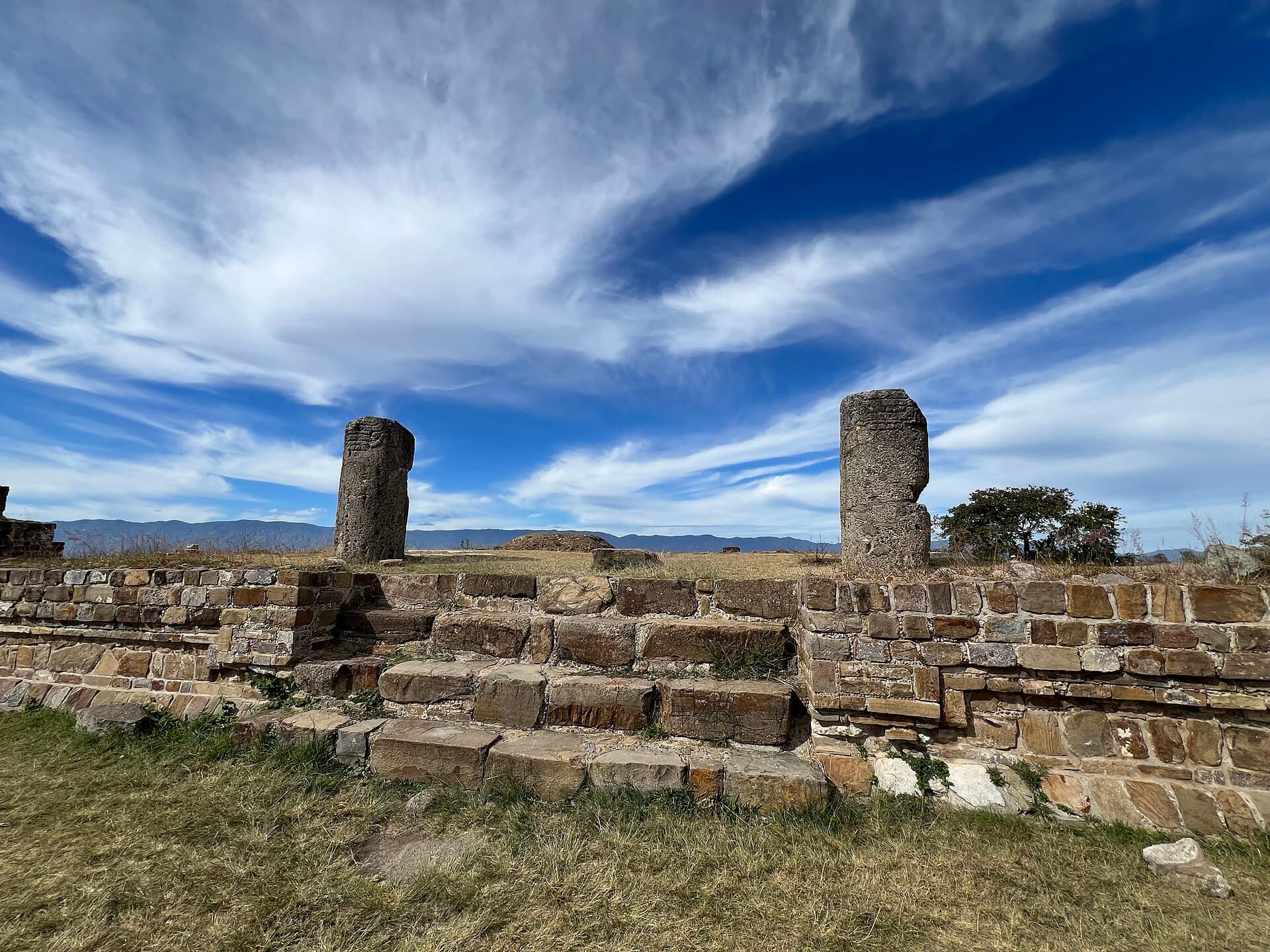 Zwei Steinsäulen und die Reste einer Plattform in Monte Albán unter zarten Wolken.