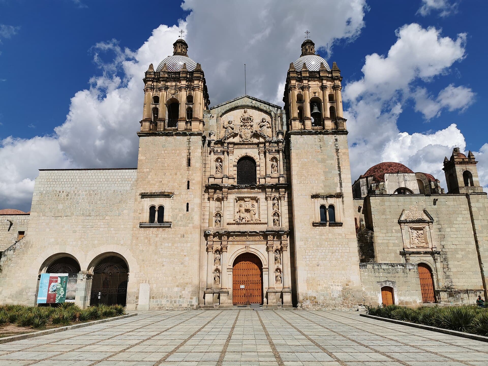 Die verzierte Steinfassade und die zwei Türme der Kirche Santo Domingo in Oaxaca, davor ein weiter Platz.