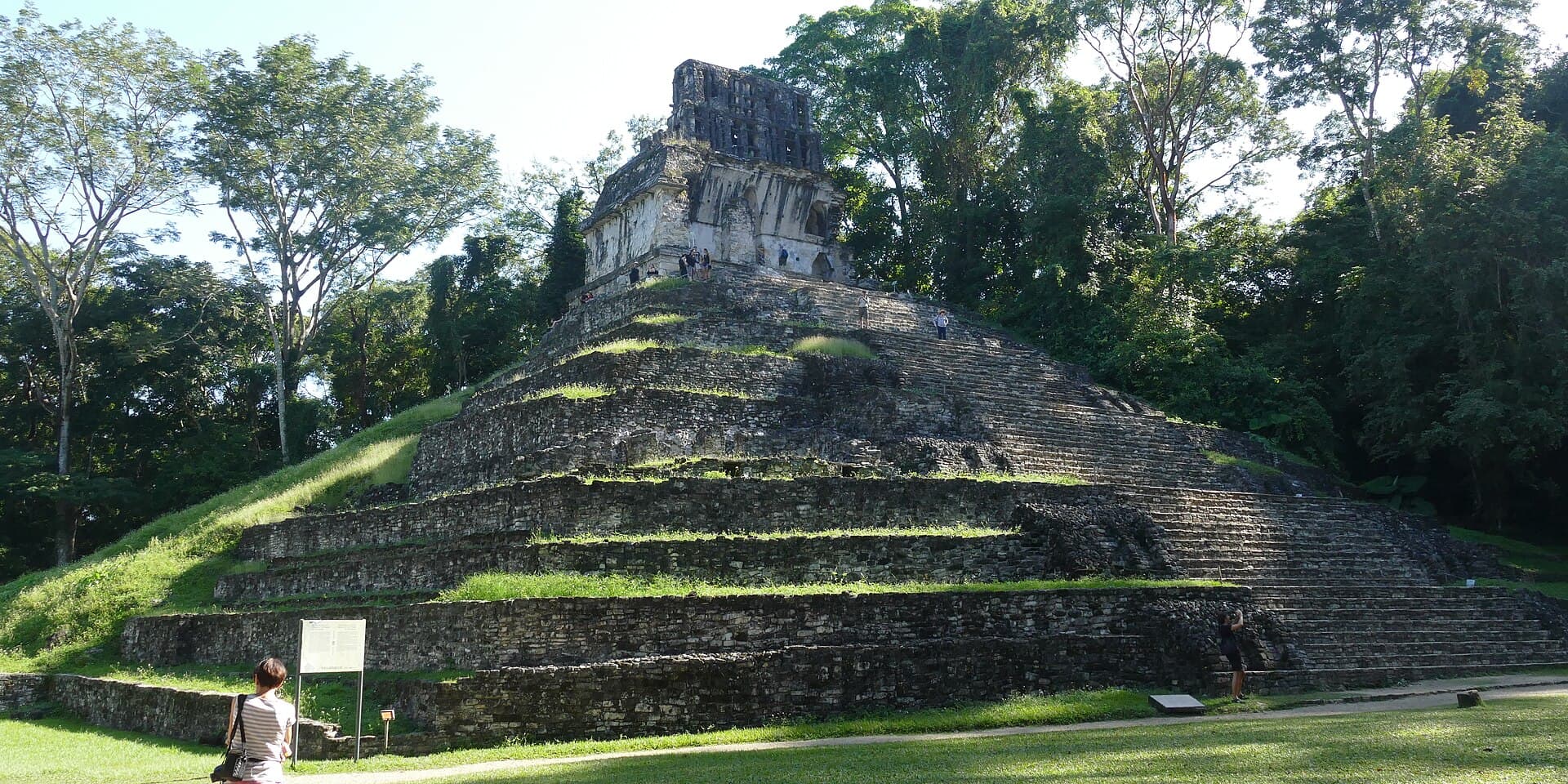 Der Kreuztempel in Palenque auf einer grasbewachsenen Stufenpyramide zwischen Waldbäumen.