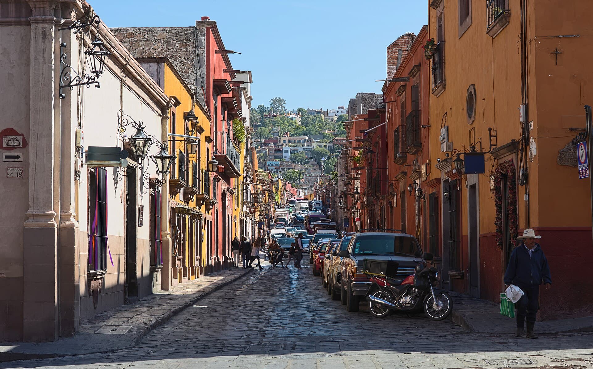 A sloping cobbled street in San Miguel de Allende between orange and yellow houses, hills on the horizon.