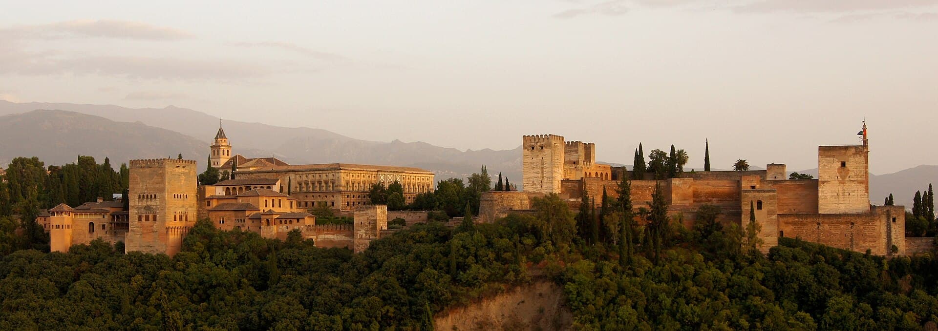 The Alhambra in Granada at evening, walls and towers on the slope with mountains behind.