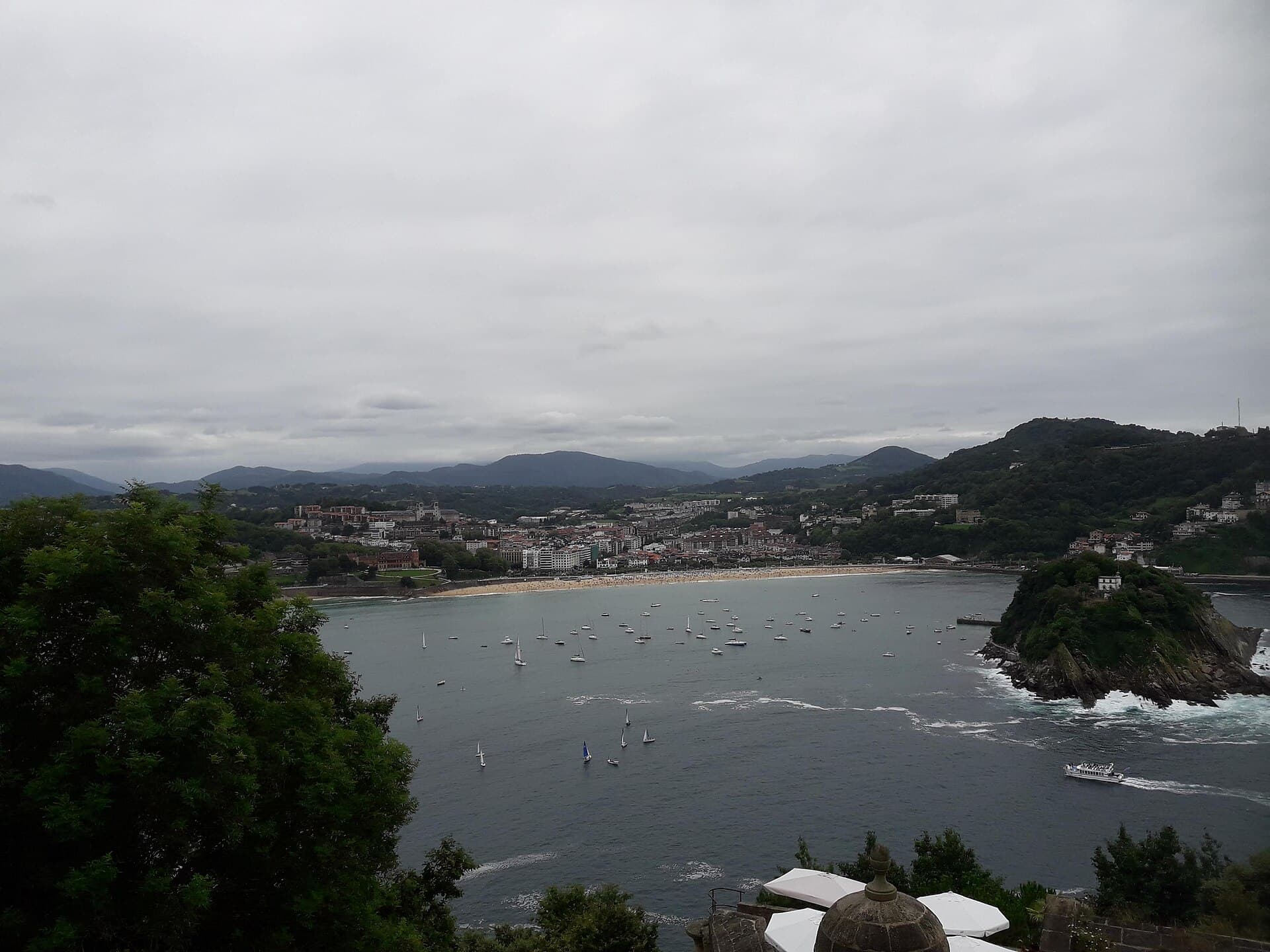 La Concha from a height, a curved beach, boats on the water and a hill on the right.