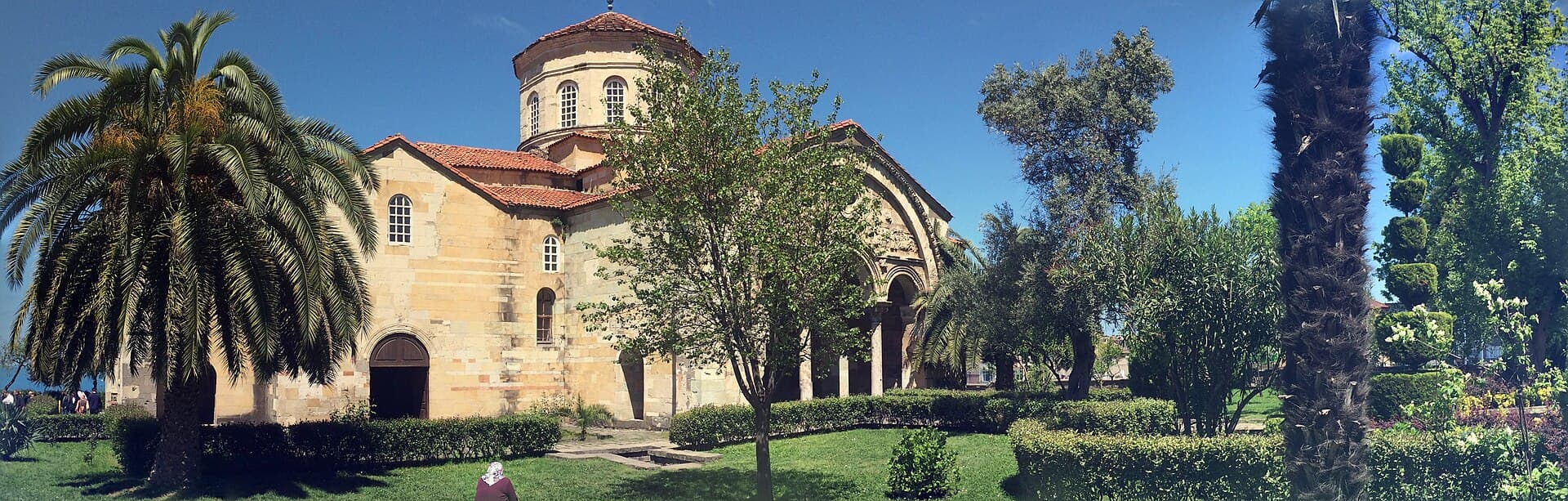 Der Steinbau der Hagia Sophia von Trabzon mit Kuppel und Bögen zwischen Bäumen und Rasen.