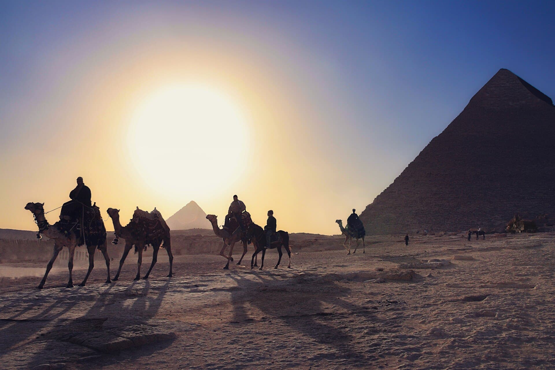Eine Kamelkarawane als Silhouette vor der Cheops-Pyramide, die Sonne tief am Himmel.