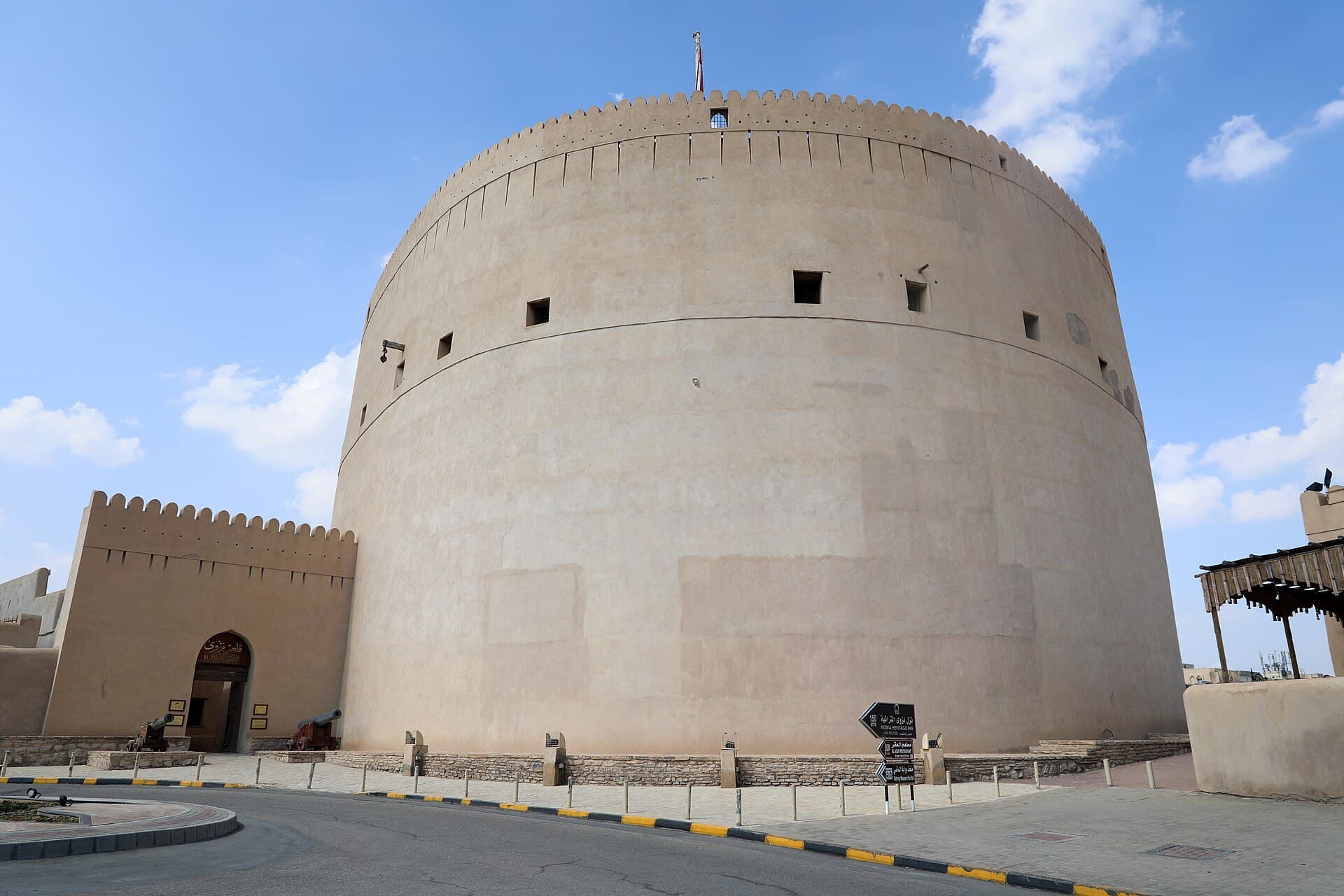 The massive round tower of Nizwa Fort from outside, beside a crenellated wall.